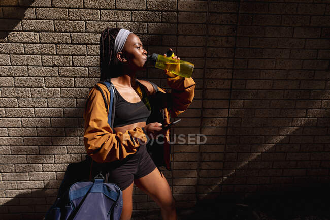 Convient femme afro-américaine avec sac de gym eau potable debout contre un mur de briques en ville. mode de vie actif urbain sain et forme physique extérieure. — Photo de stock