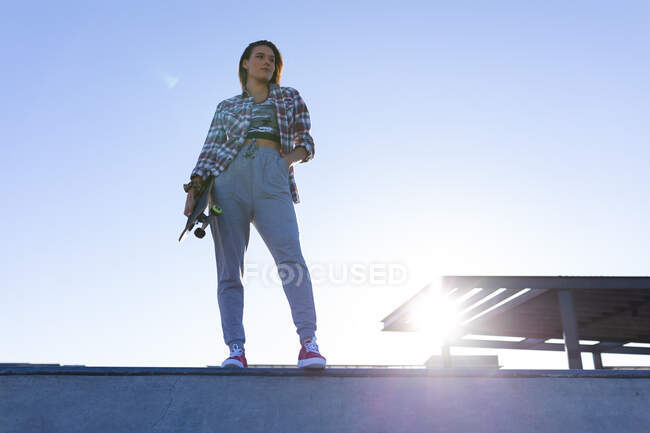 Retrato de mujer caucásica de pie en la pared con monopatín al sol. pasando el rato en un skatepark urbano en verano. - foto de stock
