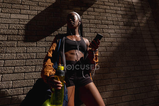 Ajuste mujer afroamericana con bolsa de gimnasio, botella de agua y teléfono inteligente de pie por la pared de ladrillo en la ciudad. estilo de vida activo urbano saludable y fitness al aire libre. - foto de stock
