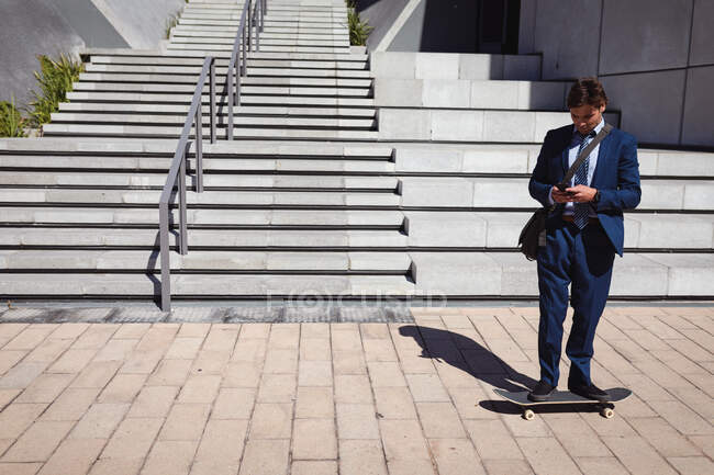 Кавказький бізнесмен стоїть на скейтборді, використовуючи смартфон на сонці. Влітку на скейтпарку.. — стокове фото