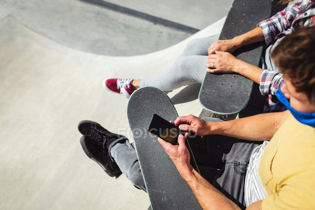 Caucasiano amigos masculinos e femininos usando máscaras faciais sentado com skates, usando smartphone. pendurado no parque de skate urbano no verão durante coronavirus covid 19 pandemia. — Fotografia de Stock