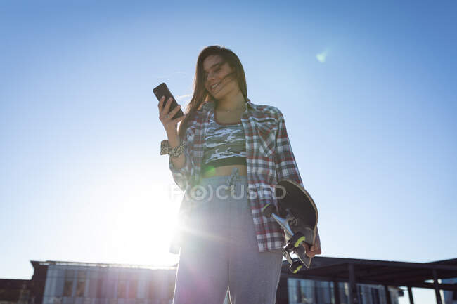 Белая женщина со скейтбордом, использующая смартфон на солнце. тусоваться в городском скейтпарке летом. — стоковое фото