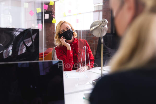 Белая женщина в маске говорит по смартфону, сидя на столе в современном офисе. гигиена и социальное дистанцирование на рабочем месте во время пандемии ковида 19. — стоковое фото