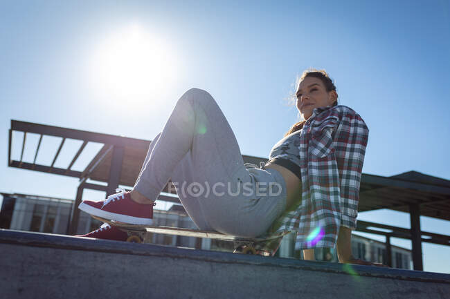 Happy caucasian woman sitting on skateboard on wall on sunny day. hanging out at urban skatepark in summer. — Stock Photo