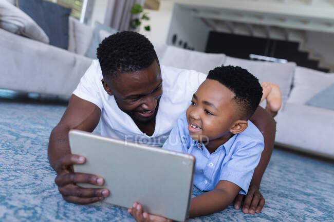Африканський американський батько і син сидять на підлозі, використовуючи планшет і посмішку. вдома в ізоляції під час карантину.. — стокове фото