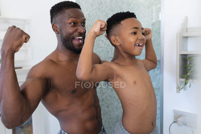 Afro-americanos pai e filho no banheiro, olhando no espelho mostrando músculos. em casa em isolamento durante o confinamento de quarentena. — Fotografia de Stock