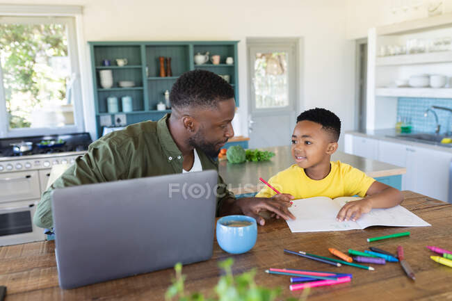Padre e hijo afroamericanos sentados en la mesa, usando un portátil y escribiendo en un cuaderno. en casa en aislamiento durante el bloqueo de cuarentena. - foto de stock