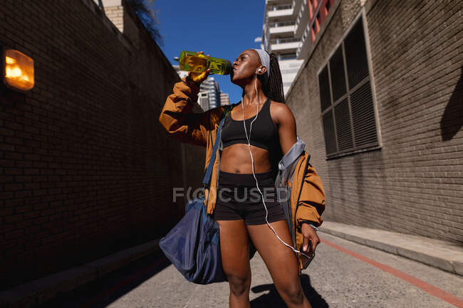 S'adapter femme afro-américaine avec sac de gym eau potable debout dans la rue dans la ville. mode de vie actif urbain sain et forme physique extérieure. — Photo de stock