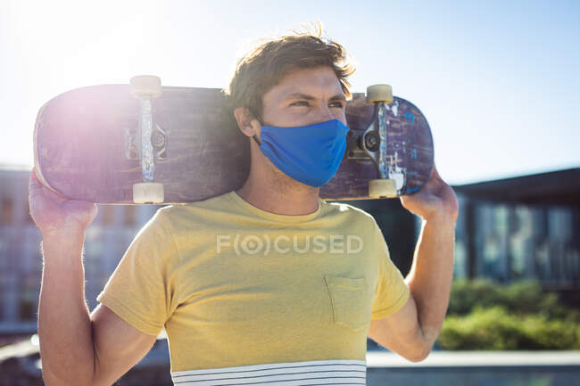 Caucasian man wearing face mask and holding skateboard. hanging out at urban skatepark in summer during coronavirus covid 19 pandemic. — Stock Photo