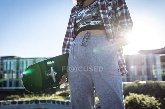 Midsection of caucasian woman standing with skateboard in the sun. hanging out at an urban skatepark in summer. — Stock Photo