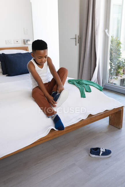 Ragazzo afroamericano seduto sul letto, che si mette le scarpe. a casa isolata durante l'isolamento in quarantena. — Foto stock