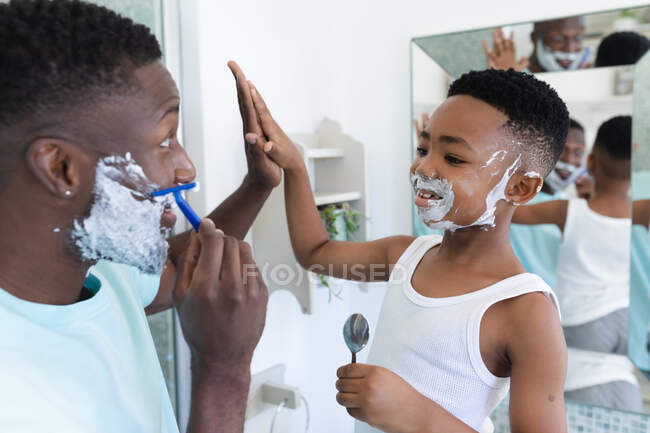 Padre e hijo afroamericanos en el baño, afeitándose juntos. en casa en aislamiento durante el bloqueo de cuarentena. - foto de stock