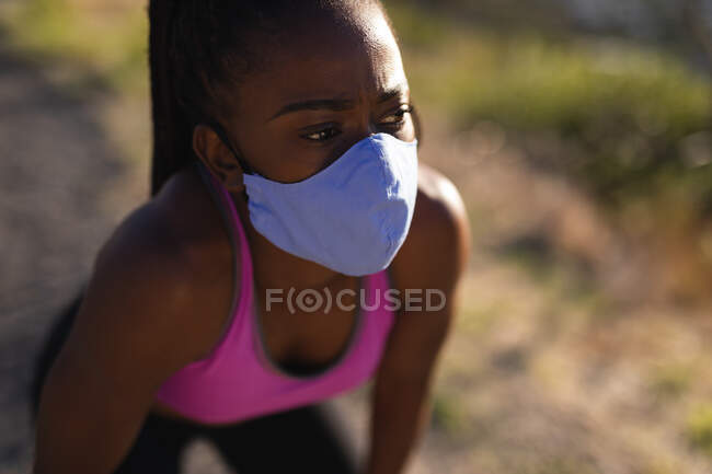 Femme afro-américaine au masque facial faisant une pause dans l'exercice à la campagne. mode de vie actif sain et forme physique en plein air pendant la covie coronavirus 19 pandémie. — Photo de stock