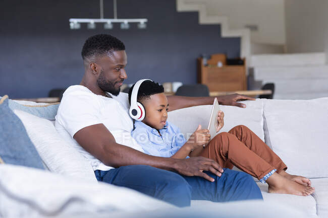 African american father and son sitting on sofa, using tablet and smiling. at home in isolation during quarantine lockdown. — Stock Photo
