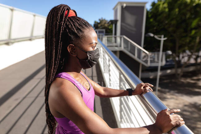 Adatto donna afro-americana in maschera facciale prendendo pausa esercizio in città. stile di vita attivo sano e fitness all'aperto durante la pandemia di coronavirus covid 19. — Foto stock