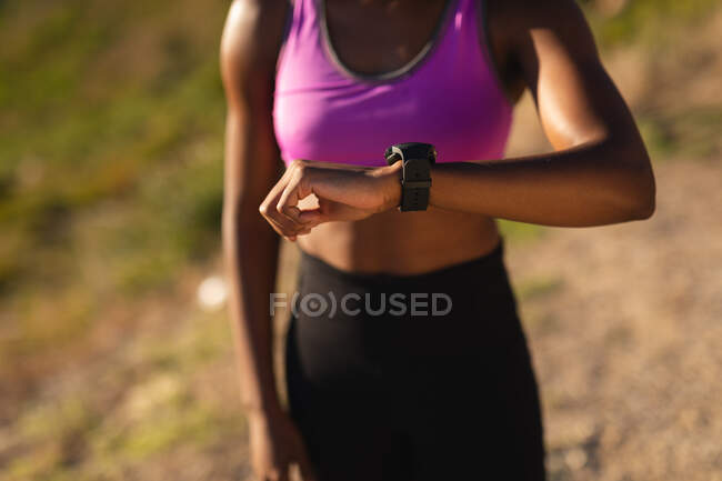 Sección media de la mujer afroamericana en forma comprobando smartwatch durante el ejercicio en el campo. estilo de vida activo saludable y fitness al aire libre. - foto de stock