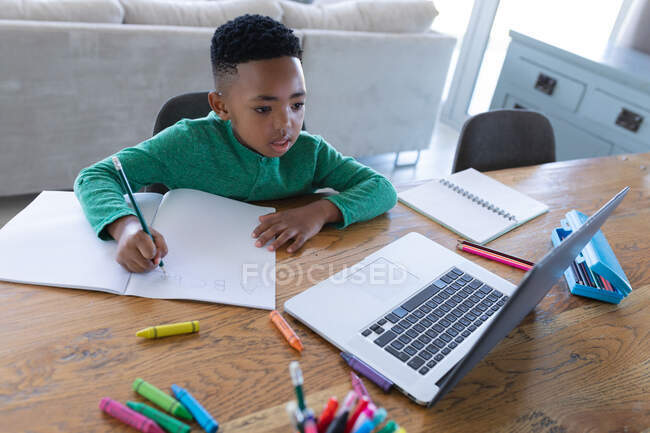 Африканський американський хлопчик в онлайн-класі, використовуючи ноутбук і письмо в блокноті. вдома в ізоляції під час карантину.. — стокове фото
