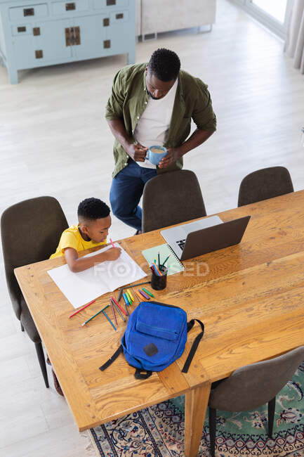 Padre e hijo afroamericanos sentados en la mesa, usando un portátil y escribiendo en un cuaderno. en casa en aislamiento durante el bloqueo de cuarentena. - foto de stock