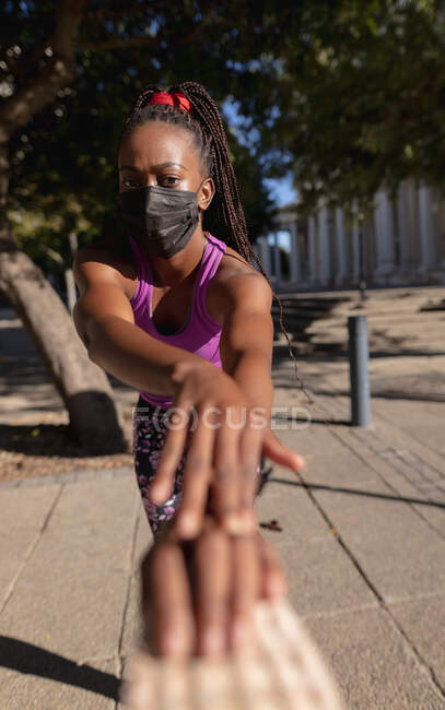 Adatto donna afro-americana in maschera che si allunga nel parco urbano. stile di vita attivo sano e fitness all'aperto durante la pandemia di coronavirus covid 19. — Foto stock