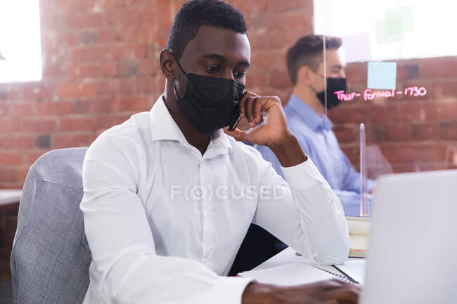 Африканский американец в маске говорит на смартфоне, используя ноутбук в офисе. гигиена и социальное дистанцирование на рабочем месте во время пандемии ковида 19. — стоковое фото