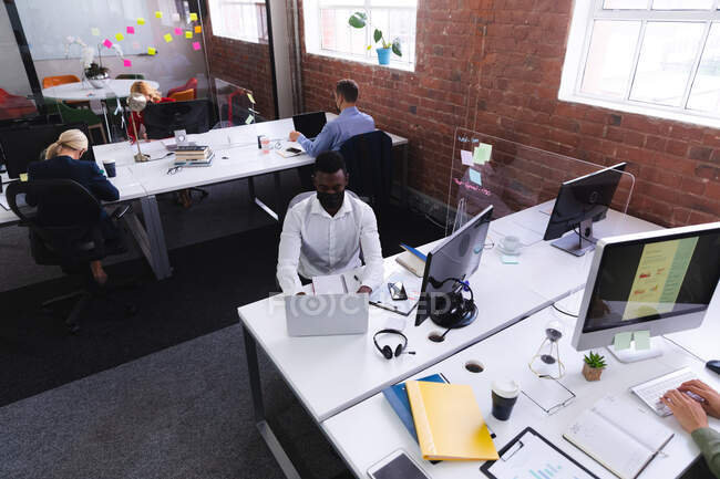 Вид сверху на африканского американца в маске для лица с помощью ноутбука в офисе. гигиена и социальное дистанцирование на рабочем месте во время пандемии ковида 19. — стоковое фото