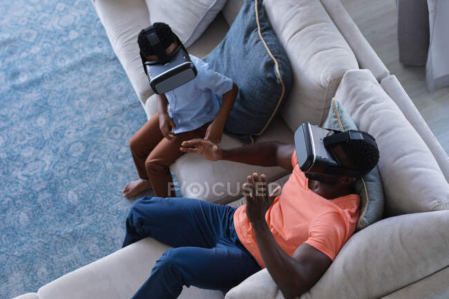 African american father and son sitting on sofa, wearing vr headset touching virtual screen. at home in isolation during quarantine lockdown. — Stock Photo