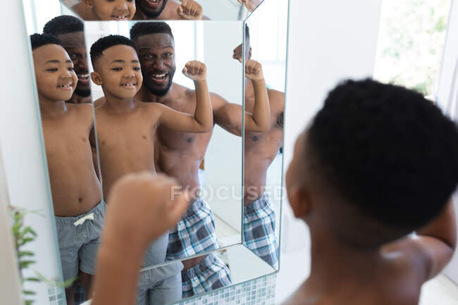 Африканский американец отец и сын в ванной комнате, смотрят в зеркало, показывая мускулы. в доме в изоляции во время карантинной изоляции. — стоковое фото