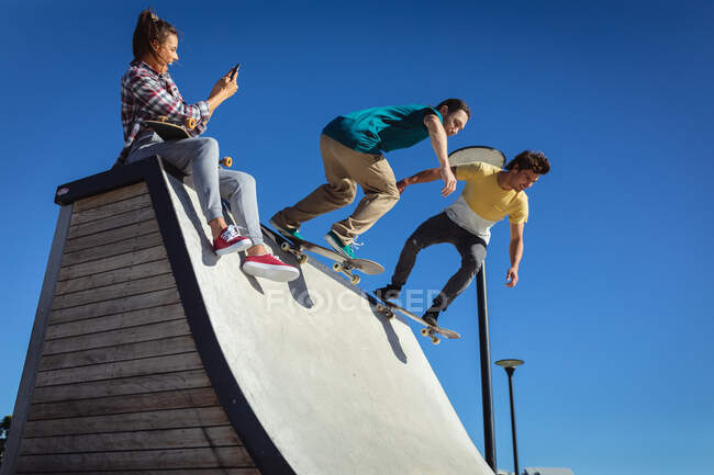 Tre felici amici caucasici, donne e uomini, che fanno skateboard al sole. uscire in estate in uno skatepark urbano. — Foto stock