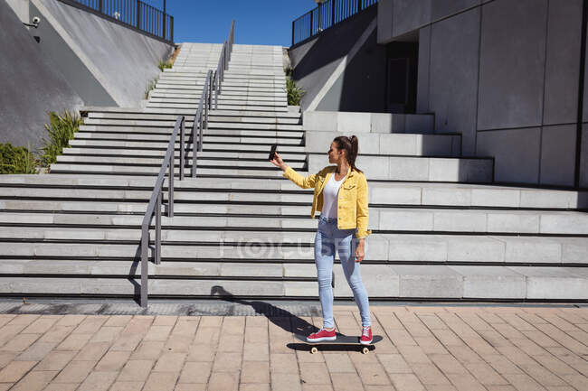 Femme caucasienne souriante debout sur le skateboard et prenant selfie à côté des escaliers. traîner à skatepark urbain en été. — Photo de stock