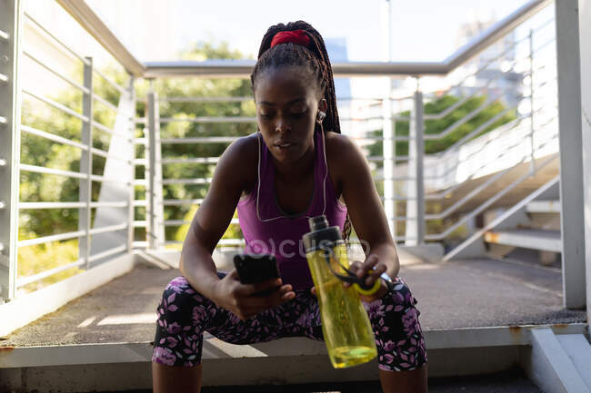 Fit african american woman sitting on steps with earphones using smartphone during exercise in city. healthy urban active lifestyle and outdoor fitness. — Stock Photo