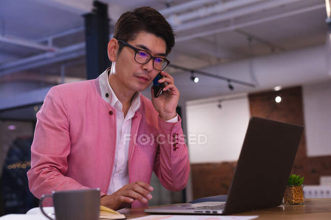 Азіат розмовляє на смартфоні, користуючись ноутбуком у сучасному офісі. бізнес, професіоналізм і офісна концепція — стокове фото