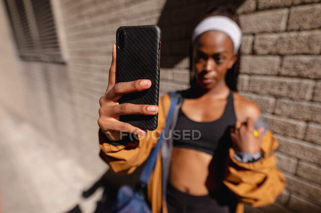 Fit afrikanisch amerikanische Frau mit Turnbeutel macht Selfie mit Smartphone steht an Ziegelmauer in der Stadt. gesunder urbaner aktiver Lebensstil und Outdoor-Fitness. — Stockfoto