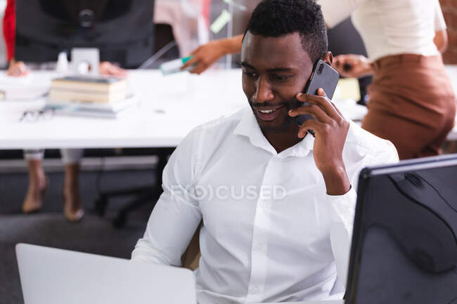 Африканський американець розмовляє на смартфоні, сидячи на столі в сучасному офісі. бізнес, професіоналізм і офісна концепція — стокове фото