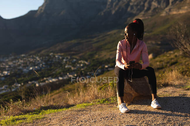 African american woman taking break in exercise outdoors, wearing earphones and holding smartphone. healthy active lifestyle and outdoor fitness. — Stock Photo