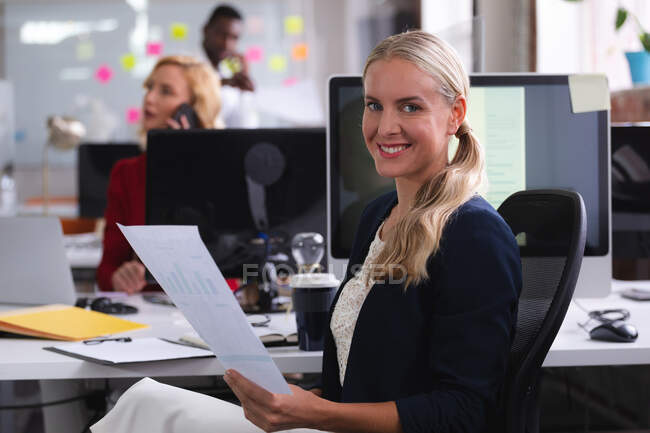 Porträt einer kaukasischen Frau, die ein Dokument lächelnd auf ihrem Schreibtisch im modernen Büro hält. Business, Professionalität und Bürokonzept — Stockfoto