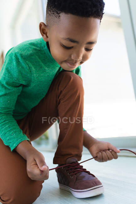 Niño afroamericano sentado en el suelo, atando cordones. en casa en aislamiento durante el bloqueo de cuarentena. - foto de stock