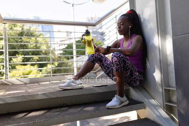 Fit Afroamerikanerin sitzt auf Stufen und überprüft Smartwatch während des Trainings in der Stadt. gesunder urbaner aktiver Lebensstil und Outdoor-Fitness. — Stockfoto
