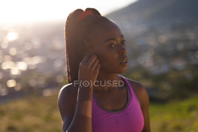 S'adapter femme afro-américaine mettre des écouteurs sans fil, prendre une pause dans l'exercice à l'extérieur. mode de vie actif sain et forme physique extérieure. — Photo de stock