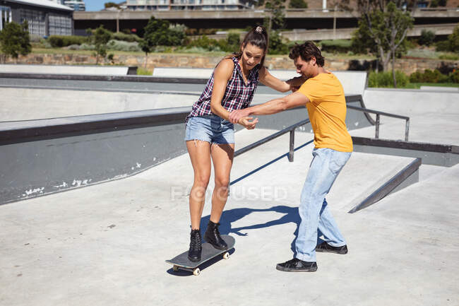 Hombre caucásico enseñando a la mujer a patinar en un día soleado. pasando el rato en skatepark urbano en verano. - foto de stock