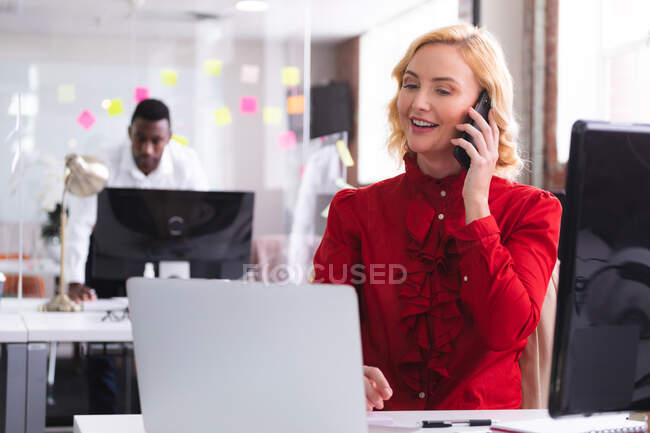 Mulher caucasiana falando no smartphone enquanto usa laptop sentado em sua mesa no escritório moderno. negócio, profissionalismo e conceito de escritório — Fotografia de Stock