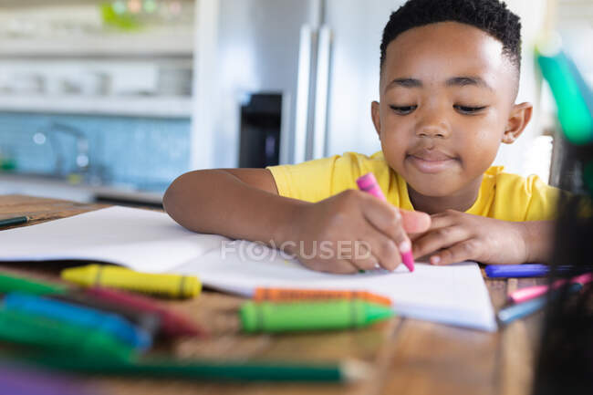 Niño afroamericano sentado en la mesa, escribiendo en cuaderno. en casa en aislamiento durante el bloqueo de cuarentena. - foto de stock
