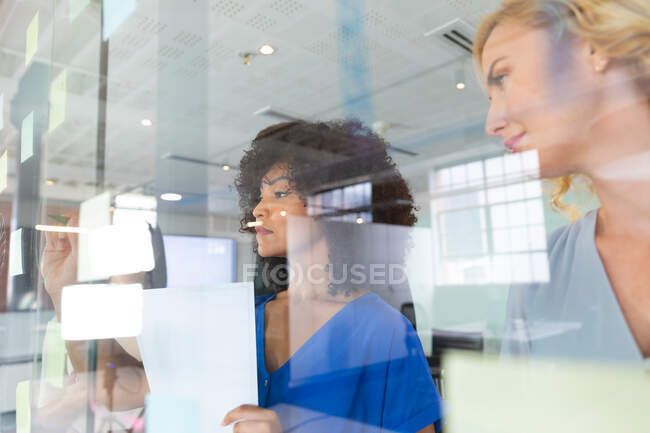 Дві різні жінки-офісні колеги пишуть на скляній дошці в сучасному офісі. бізнес, професіоналізм, концепція офісної та командної роботи — стокове фото