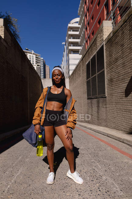 Donna afroamericana in forma sorridente con borsa da palestra in piedi in strada in città. stile di vita attivo urbano sano e fitness all'aperto. — Foto stock