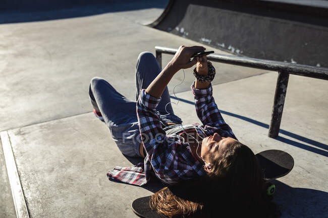 Mujer caucásica acostada en la escalera con monopatín, usando smartphone al sol. pasando el rato en un skatepark urbano en verano. - foto de stock