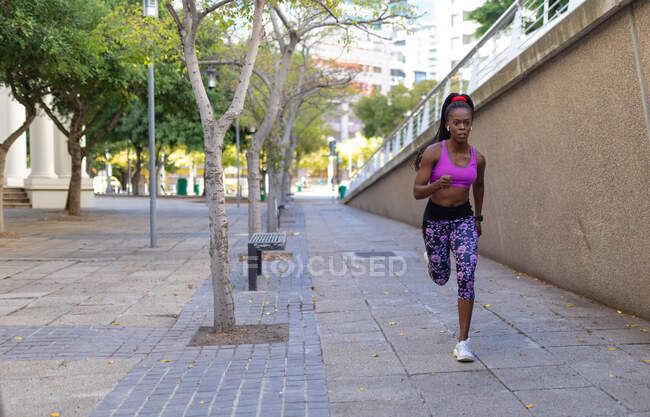 S'adapter femme afro-américaine en cours d'exécution portant des écouteurs sans fil exercice en ville. mode de vie actif urbain sain et forme physique extérieure. — Photo de stock
