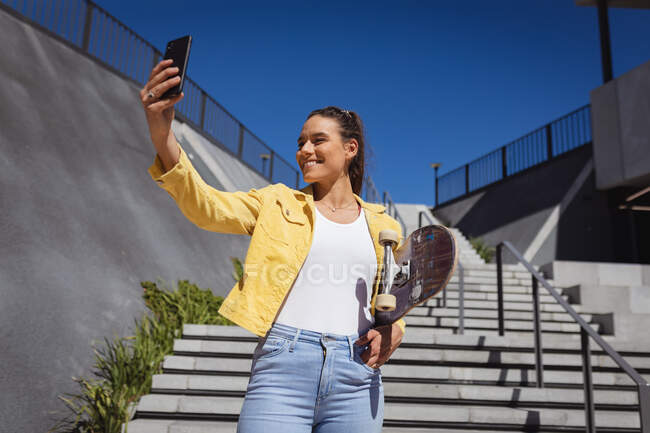 Mujer caucásica sonriente sosteniendo monopatín y tomando selfie al lado de las escaleras. pasando el rato en skatepark urbano en verano. - foto de stock