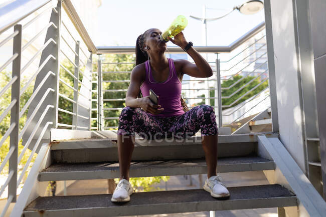 Fit african american woman sitting on steps drinking water taking break during exercise in city. healthy urban active lifestyle and outdoor fitness. — Stock Photo