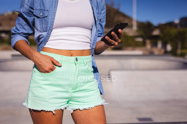 Середина женщины, стоящей и использующей смартфон. тусоваться в городском скейтпарке летом. — стоковое фото
