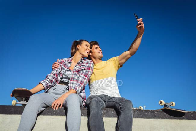 Glückliche kaukasische Frau und Mann, die mit Skateboards an der Wand sitzen und Selfies in der Sonne machen. Im Sommer im städtischen Skatepark abhängen. — Stockfoto
