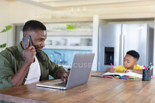 Afro-americano pai e filho sentado à mesa, usando laptop falando no smartphone. em casa em isolamento durante o confinamento de quarentena. — Fotografia de Stock